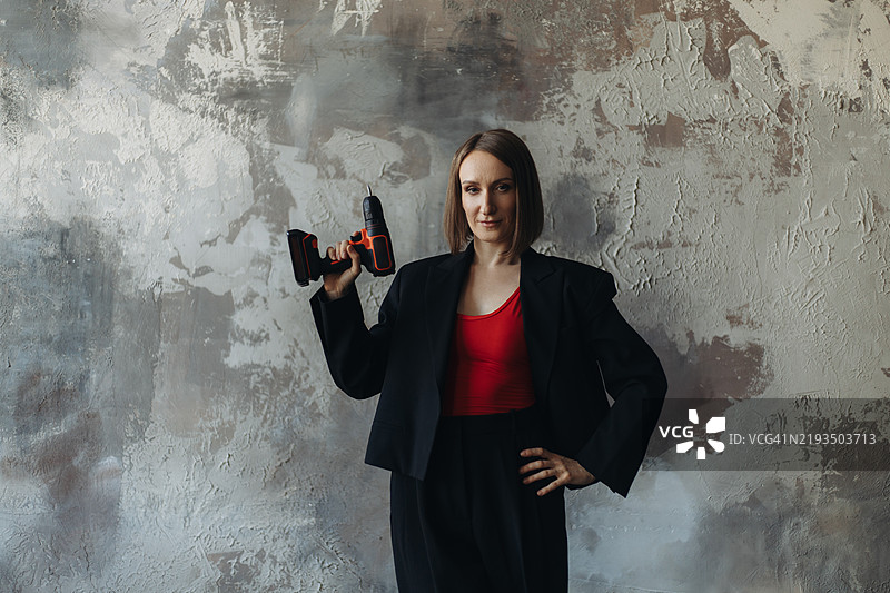 年轻女性穿着黑色夹克和红色上衣，手持电动螺丝刀摆姿势。图片素材