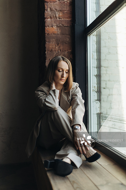 年轻女性穿着浅色西装坐在砖墙旁的窗台上。图片素材