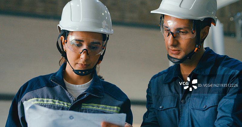 两名年轻工程师穿着安全装备，正在机器人工厂内分析笔记本电脑和文件上的数据，专注于精确和创新。机器人工业工厂。图片素材