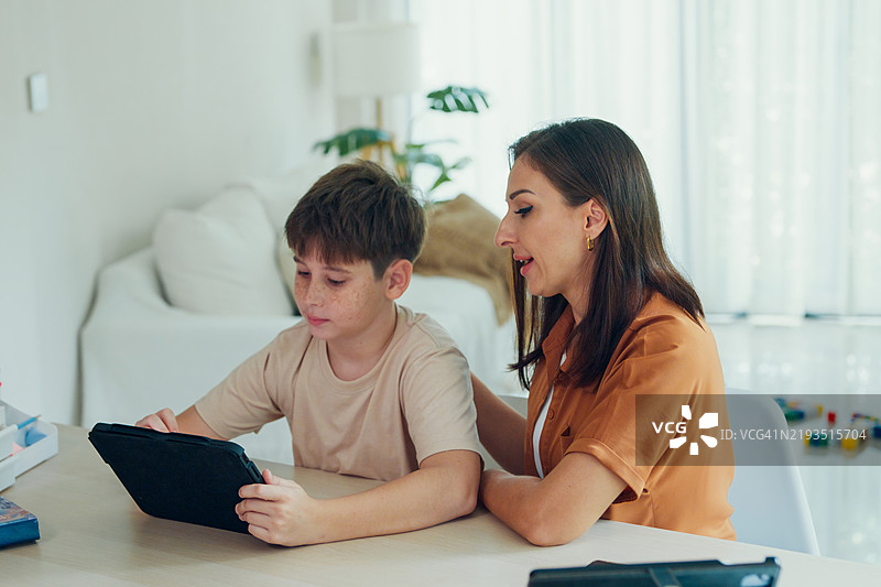 年轻母亲在明亮的家居环境中帮助儿子使用平板电脑进行在线学习，提供指导和支持，他们坐在桌子旁。返校概念。图片素材