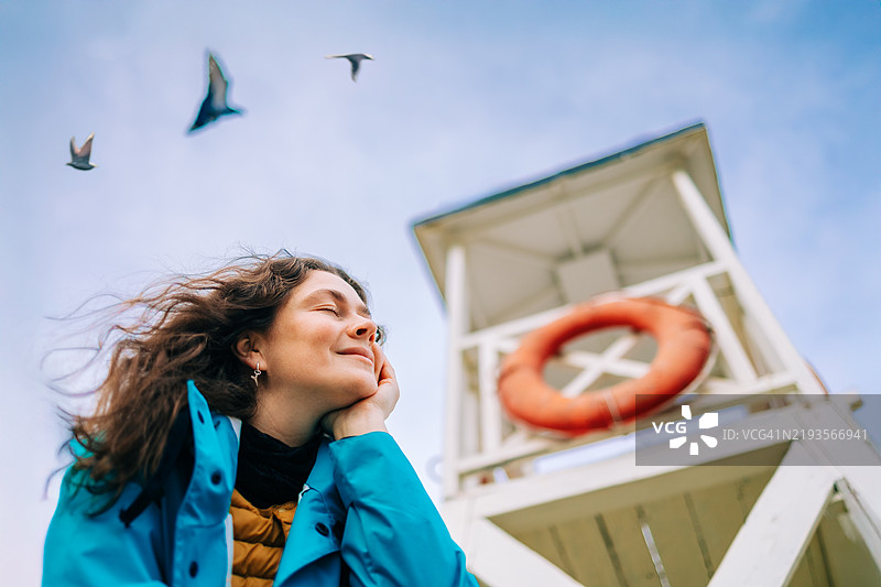 女人在海滩上享受清新的海风，身旁有救生塔和飞翔的鸟儿图片素材