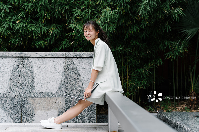 一位年轻的亚洲白领女性坐在公园长椅上，面带微笑看向镜头。图片素材
