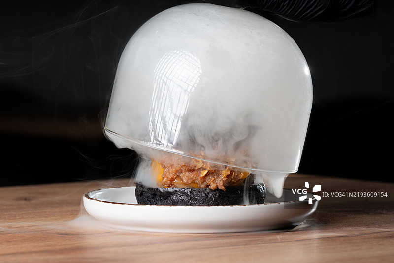 厨师举起玻璃罩，揭示冒烟的美味黑色汉堡图片素材