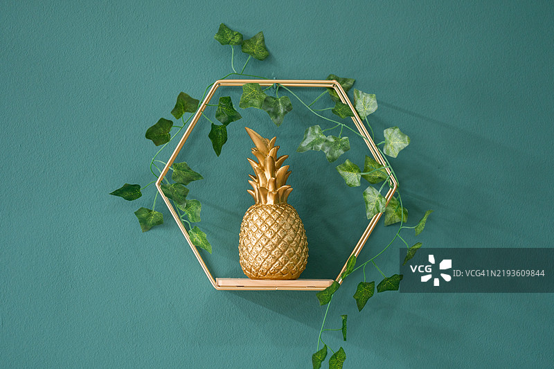 金色菠萝装饰在六角形架子上，旁边有常春藤，背景是青绿色墙壁。图片素材