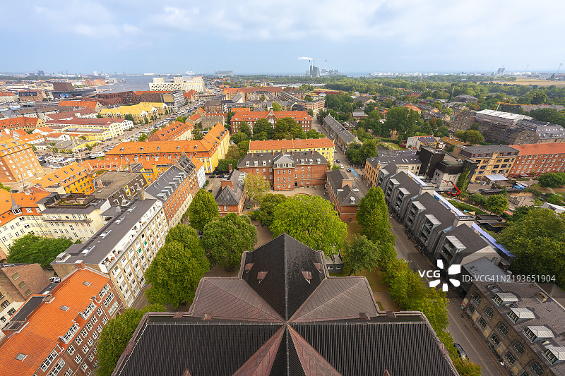 从救世主教堂顶部俯瞰哥本哈根的克里斯蒂安港社区，展现了五彩斑斓的屋顶、住宅建筑、郁郁葱葱的树木以及远处的海滨，天空部分多云。图片素材