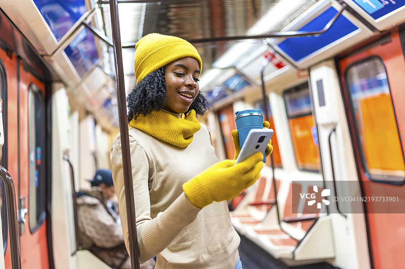 微笑的黑人女性穿着黄色冬季服装，在地铁车厢内使用手机并手持可重复使用的咖啡杯。图片素材
