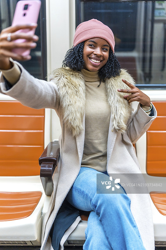 快乐的年轻女性在公共交通上自拍，微笑着用手指做出和平手势。图片素材