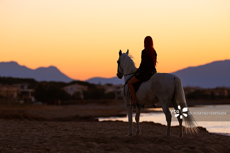 迷人的剪影，一位女性骑着她的白马在海滩上迎着日落骑行，靠近大海。图片素材