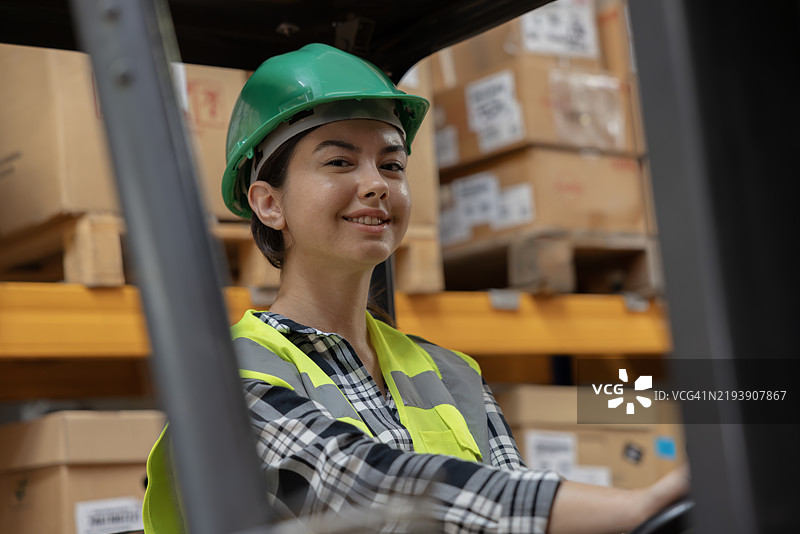 高效的仓库操作：女性叉车司机在配送仓库中摆姿势并驾驶工作叉车。图片素材