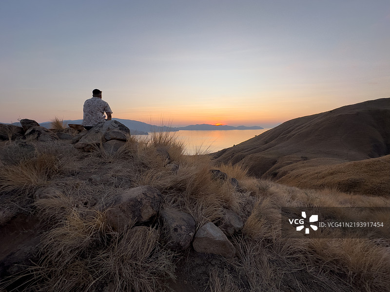 一个男人背对着海洋坐着，观看印度尼西亚科莫多国家公园拉瓦达拉特吉利岛上的日落。图片素材