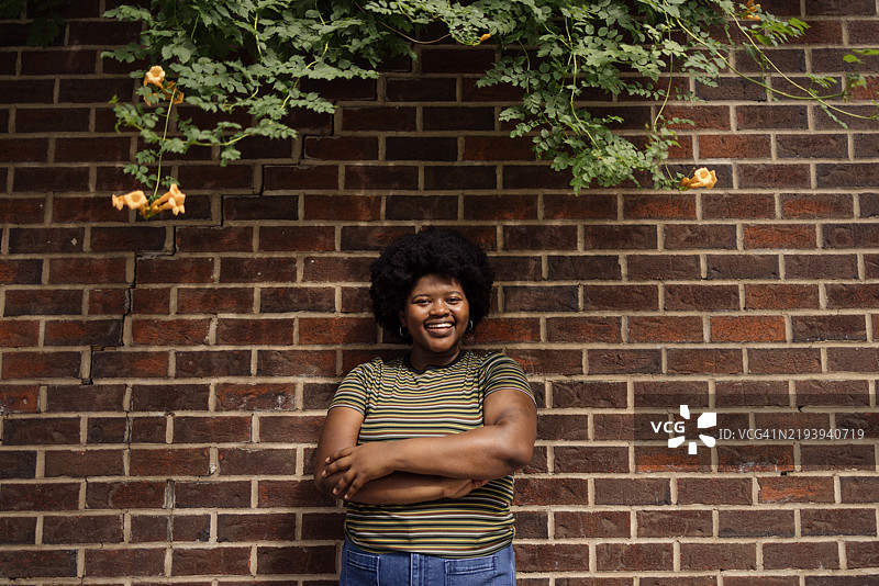 微笑的女性站在砖墙前，背景是绿色植物。图片素材