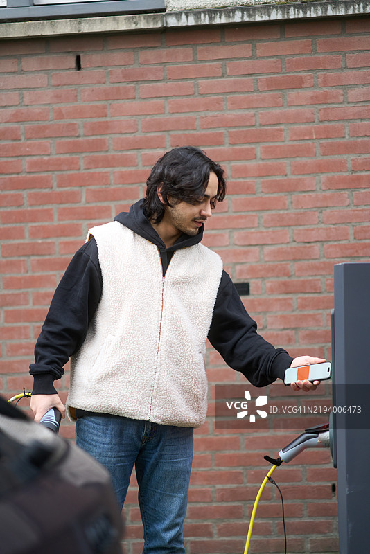 年轻男子正在为电动车充电，并使用智能手机付款图片素材