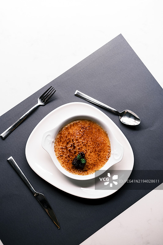 经典焦糖布丁的俯视图，上面有焦糖化的糖壳和浆果装饰，优雅地放在白色烤碗中，搭配现代黑白餐桌布置和银器。图片素材