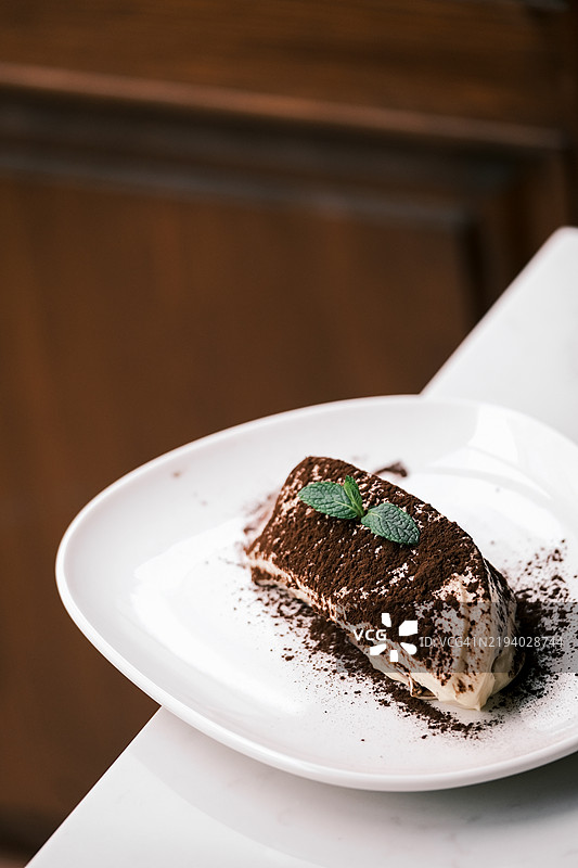 经典提拉米苏甜点特写，撒上可可粉和薄荷装饰，优雅地摆放在温馨的意大利餐厅环境中的白色盘子上，精致餐饮与美食糕点概念图片素材
