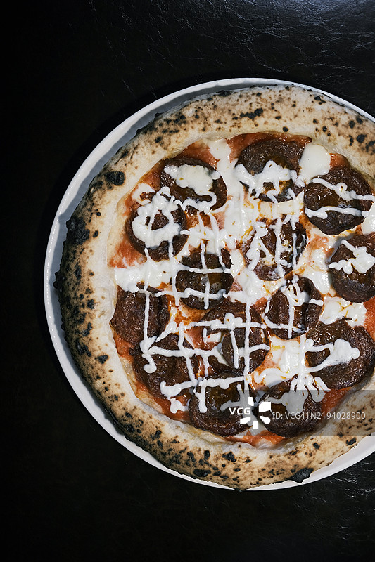 从上方俯视美味的意大利披萨，酥脆的外壳，香肠，融化的奶酪，淋上白色酱汁，放在古朴的盘子里，置于昏暗的餐厅桌上，真实的木火披萨体验图片素材