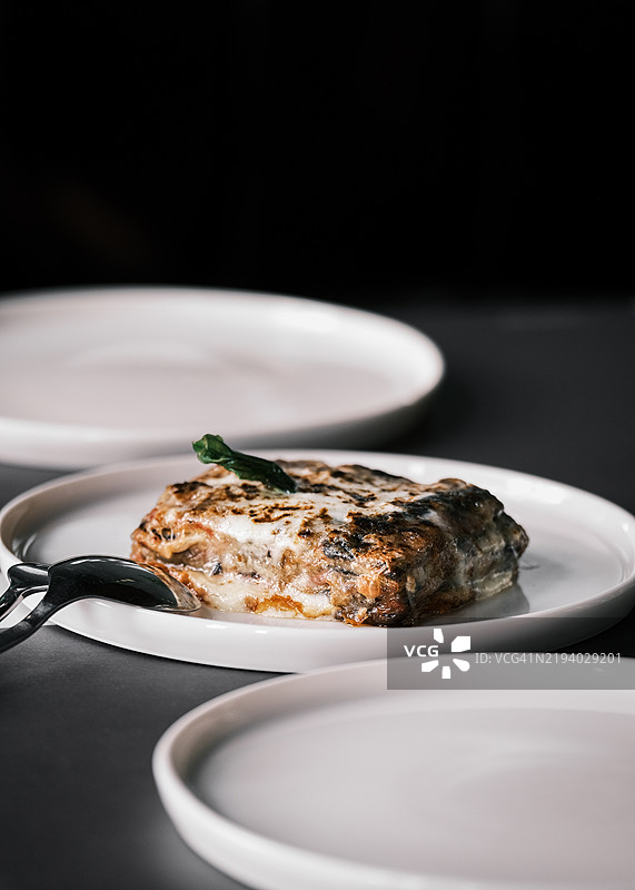 茄子帕尔马干酪，白色盘子盛装，上面点缀着罗勒，层叠着马苏里拉奶酪和番茄酱，经典意大利菜肴，餐厅用餐环境，优雅的摆盘和柔和的灯光。图片素材