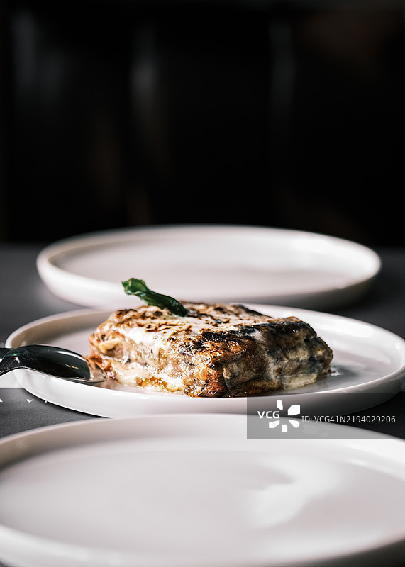 茄子帕尔马干酪，白色盘子盛装，上面点缀罗勒，层叠着马苏里拉奶酪和番茄酱，经典意大利菜肴，餐厅用餐环境，优雅的摆盘和柔和的灯光。图片素材