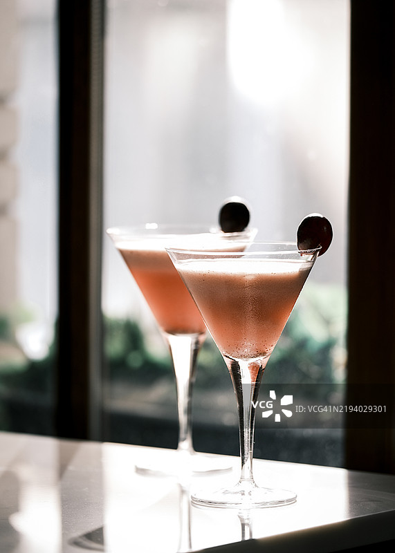 优雅的粉色鸡尾酒盛在马提尼酒杯中，点缀着深色樱桃，摆放在意大利餐厅阳光明媚的窗边，展现出精致的调酒艺术，时尚的酒吧氛围，清爽的晚间饮品图片素材