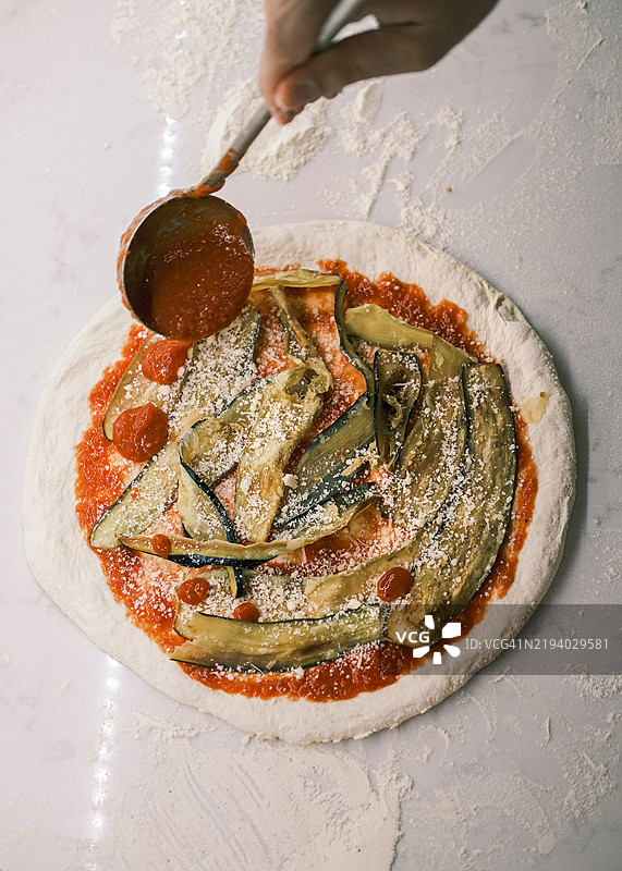 厨师在专业比萨厨房中，特写镜头展示他在生比萨面团上添加额外的番茄酱、烤茄子和磨碎的奶酪，传统意大利比萨的制作过程。图片素材