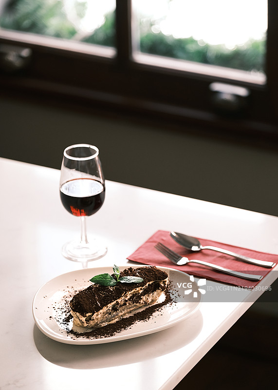 经典意大利提拉米苏甜点，配以可可和薄荷，优雅地与一杯红酒搭配，摆放在一家正宗意大利餐厅的白色桌子上。图片素材