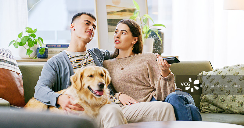 放松的狗和一对情侣在家看电视，增进彼此的联系或家庭时光。年轻的男女与金毛猎犬小狗一起坐在沙发上，在线观看电影或节目。图片素材