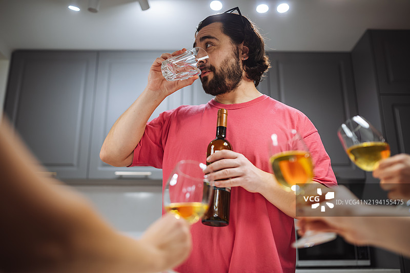 穿着粉色衬衫的男人正在喝水，手里拿着一瓶葡萄酒，前景中有几杯葡萄酒。适合社交聚会和派对的图片。图片素材