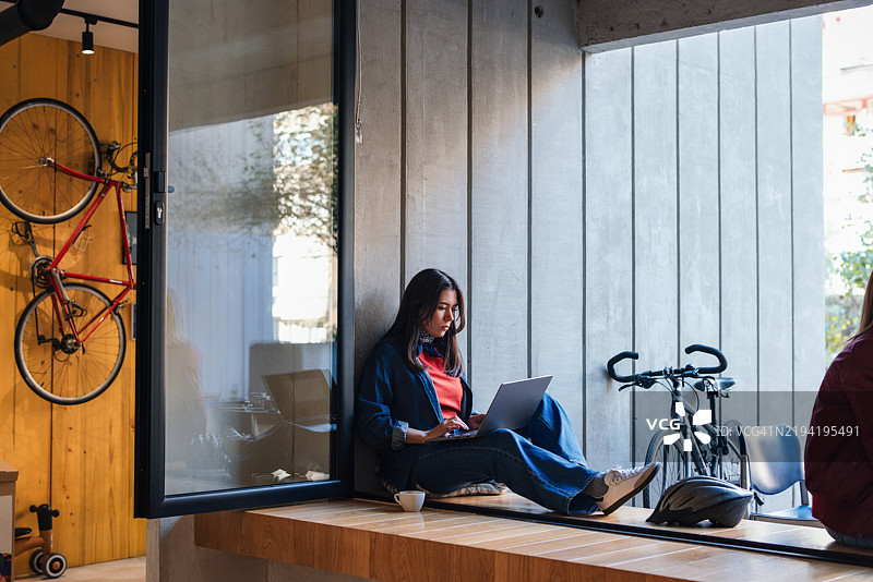 年轻女性在咖啡馆窗边用笔记本电脑工作，旁边有一辆自行车图片素材