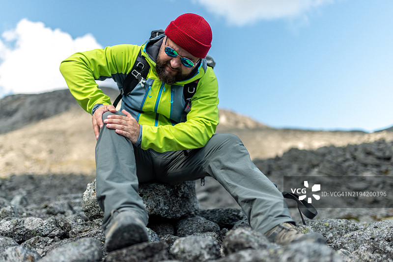户外徒步者痛苦地捂着膝盖，背景是崎岖的山脉图片素材