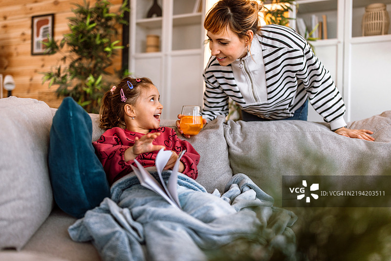 一个小女孩躺在沙发上，盖着毯子，正在看书，而她的妈妈则给她送来新鲜的橙汁。图片素材
