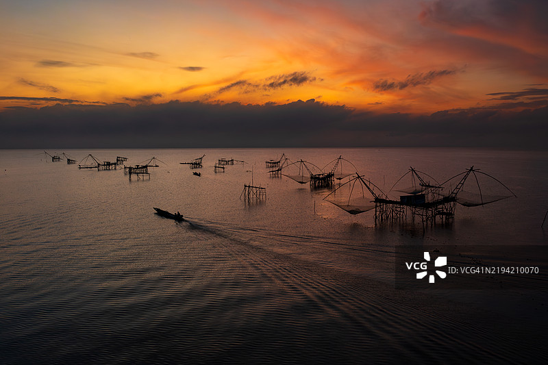 在泰国帕塔尔龙的帕克普拉河上，日出时分，一只船的剪影驶过传统的渔网和平台。图片素材