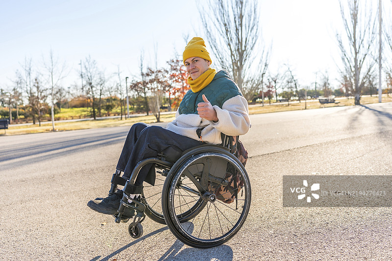阳光明媚的日子里，一位年轻的残疾男子在公园里坐在轮椅上竖起大拇指。图片素材