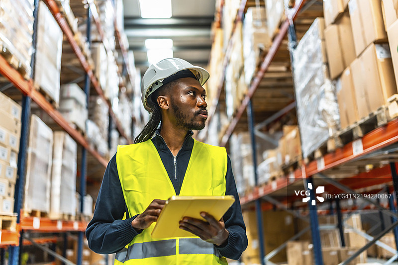 身穿保护头盔和黄色反光背心的非洲工人在仓库中使用数字平板电脑进行库存盘点图片素材