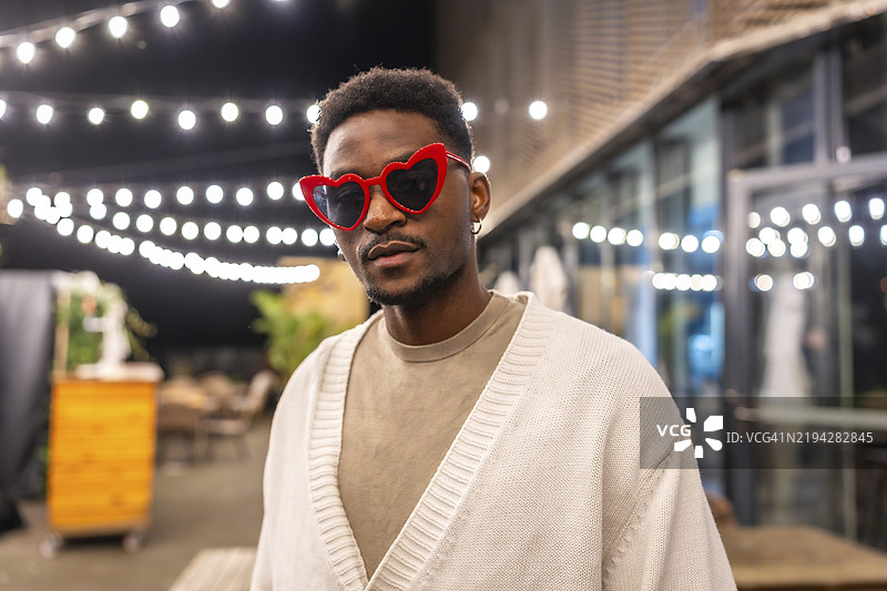 现代酷炫的非洲男子，戴着心形太阳镜，站在夜晚的酒吧露台上图片素材