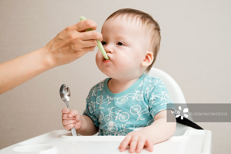 婴儿男孩坐在高脚椅上吃无味的土豆泥，母亲正在喂他，手里拿着勺子为他准备蔬菜午餐，宝宝正在断奶，第一次吃固体食物。图片素材