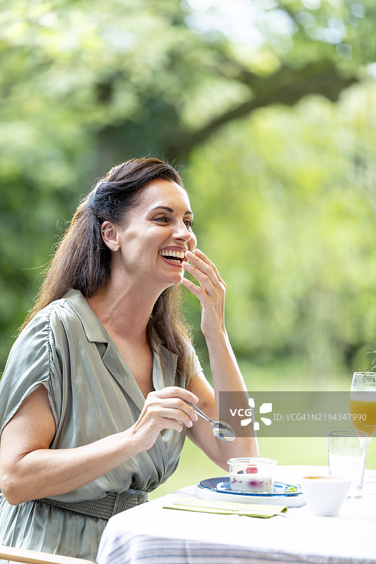 一位长发黑发的成熟的女性在阳光明媚的夏日早晨，在花园里享用健康早餐，显得非常开心并且在大笑。图片素材