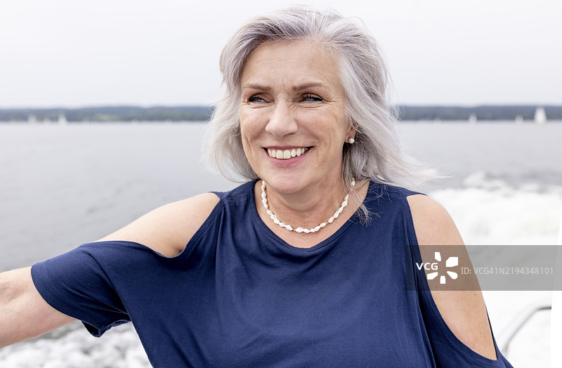 快乐的老年女性，长长的灰发，穿着蓝色连衣裙，在湖上的摩托艇上享受时光。图片素材