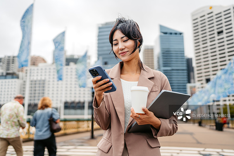 年轻的亚洲女商人在澳大利亚悉尼达令港一边喝外带咖啡一边使用手机，同时手里还拿着一台笔记本电脑。图片素材