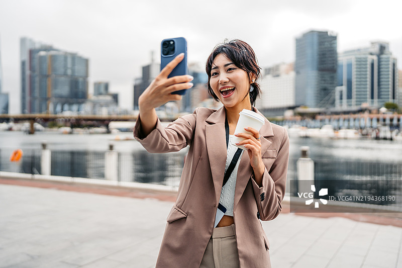 年轻的亚洲女商人在澳大利亚悉尼达令港一边喝外带咖啡一边用手机自拍图片素材