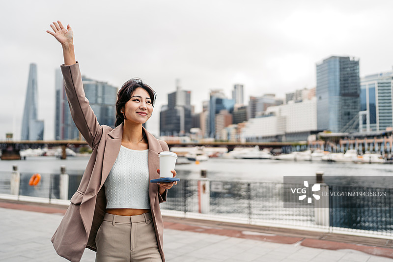 年轻的亚洲女商人在澳大利亚悉尼达令港边喝着外带咖啡招手叫出租车图片素材