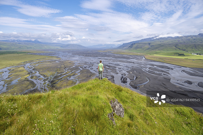 年轻男子在山丘上远足，俯瞰冲积平原和蜿蜒的河流，地点：Dímonarhellir，南部地区，冰岛，欧洲图片素材