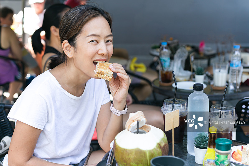 快乐的亚洲女性在当地咖啡馆户外享用金枪鱼三明治图片素材