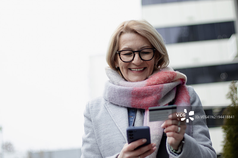 一位面带微笑的成熟金发女商人在办公楼前戴着眼镜，使用信用卡在线支付账单。图片素材