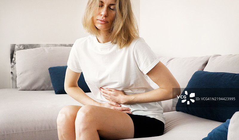 年轻的金发女性坐在家里的沙发上，感到腹部疼痛。图片素材