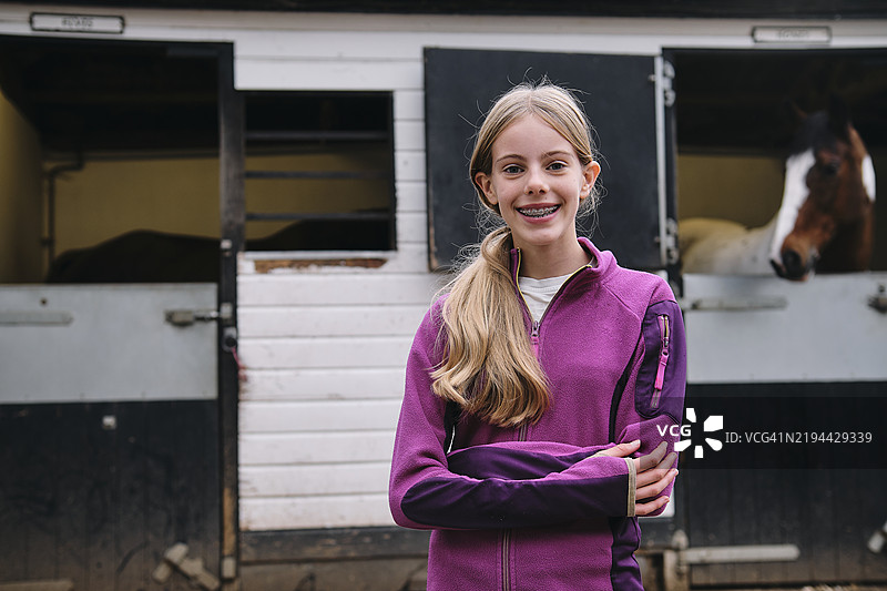 年轻骑手微笑着，双臂交叉站在马厩前。图片素材
