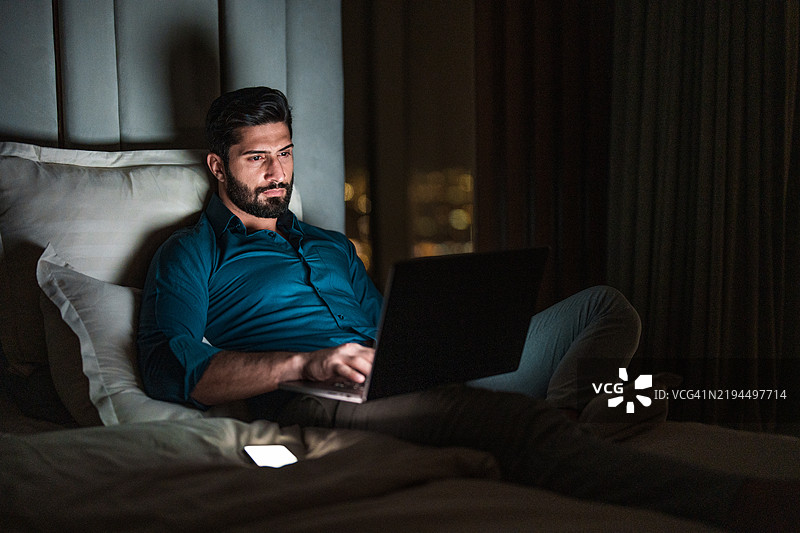 专注的年轻阿拉伯男性在昏暗的房间里使用笔记本电脑图片素材