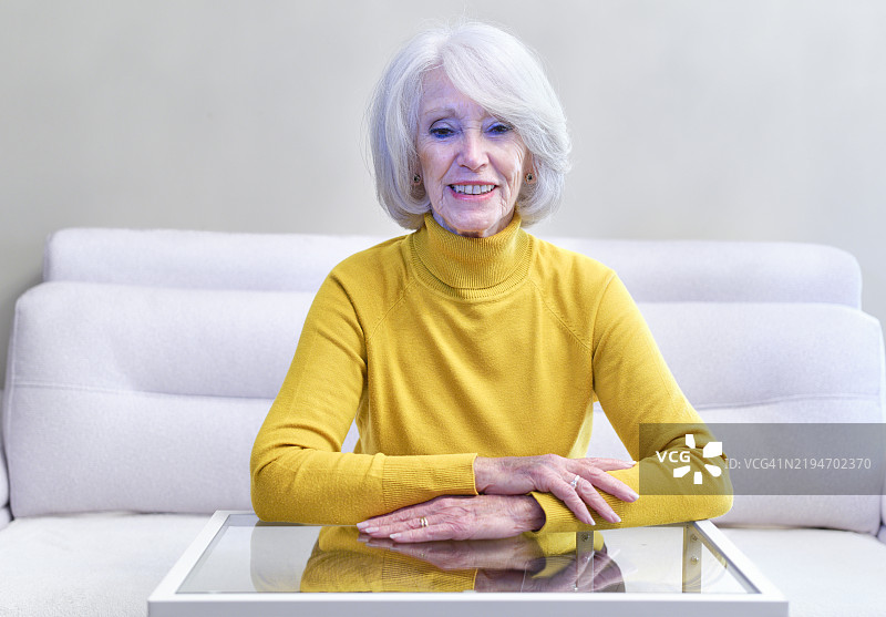 微笑的老年女性坐在家中的沙发上，双手交叠，面向镜头，背景色彩绚丽。图片素材