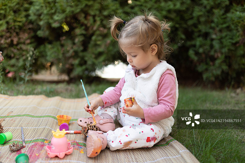 一个一岁的小女孩坐在地上，用蛋彩画给松果上色。图片素材
