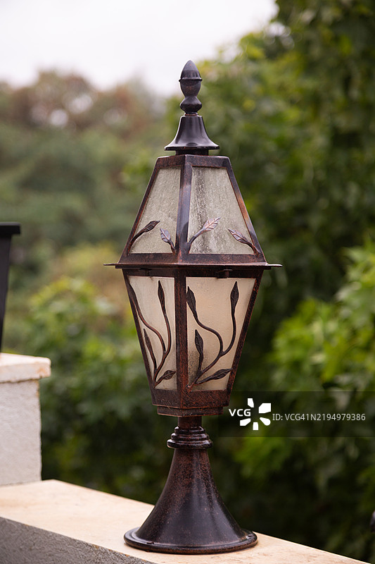 优雅的户外灯笼放置在基座上，具有乡村风格的青铜饰面和装饰性叶子图案的磨砂玻璃面板，背景是模糊的绿色树叶。图片素材