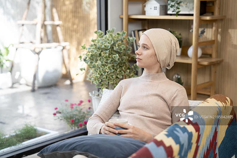 一位年轻女性在家中的沙发上与阳光相伴，正在与癌症抗争，治疗间隙享受阳光。她穿着休闲，头上裹着头巾以保持温暖。图片素材