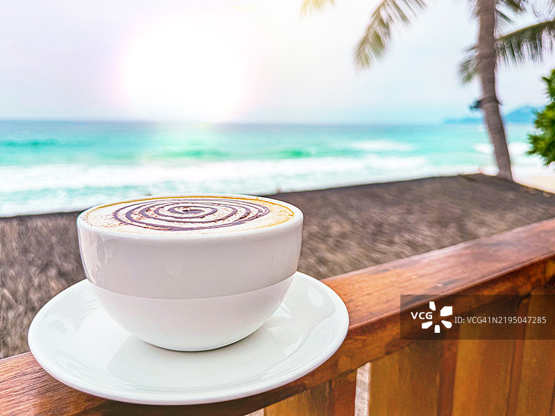 热带海滩上，白色瓷杯中的香浓咖啡卡布奇诺，背景是棕榈树和蓝色海洋，夕阳时分。图片素材